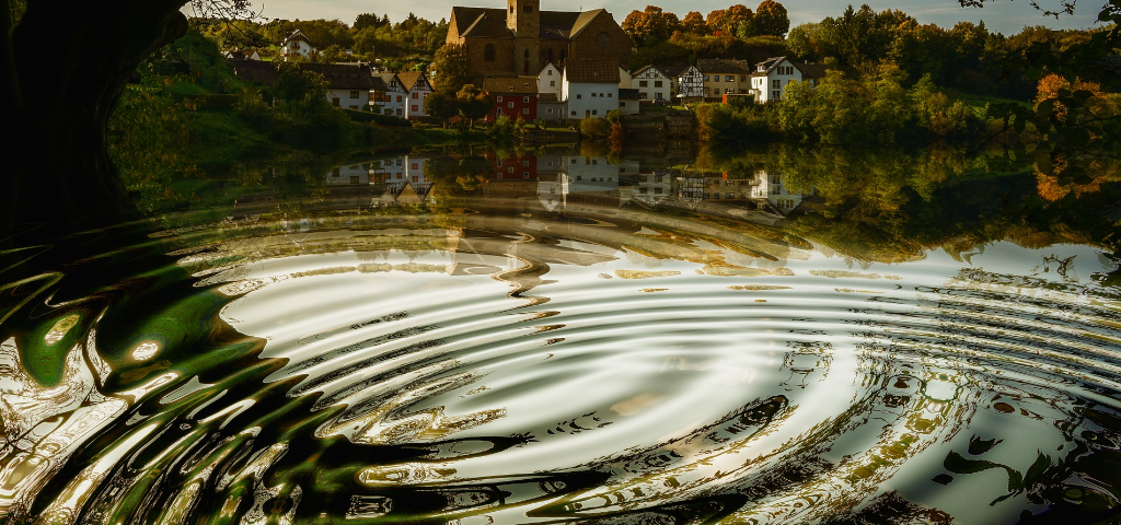 Image of lake with ripples of water stretching out underneath tree limbs and a church visible on the other side of the lake - photo by Gerd Altman on pixabay