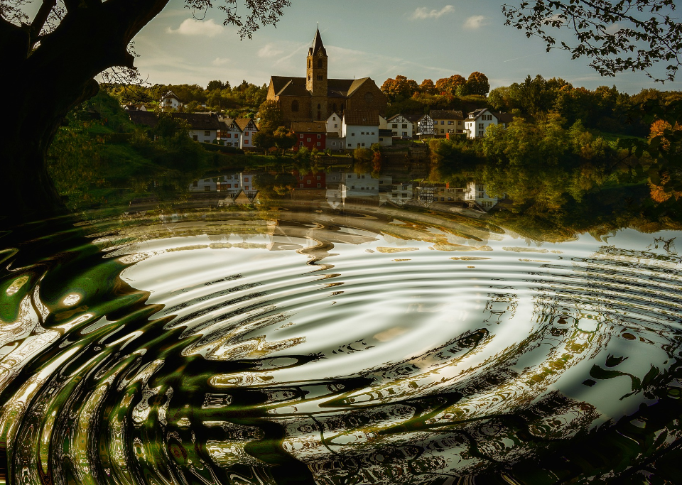 Image of lake with ripples of water stretching out underneath tree limbs and a church visible on the other side of the lake - photo by Gerd Altman on pixabay
