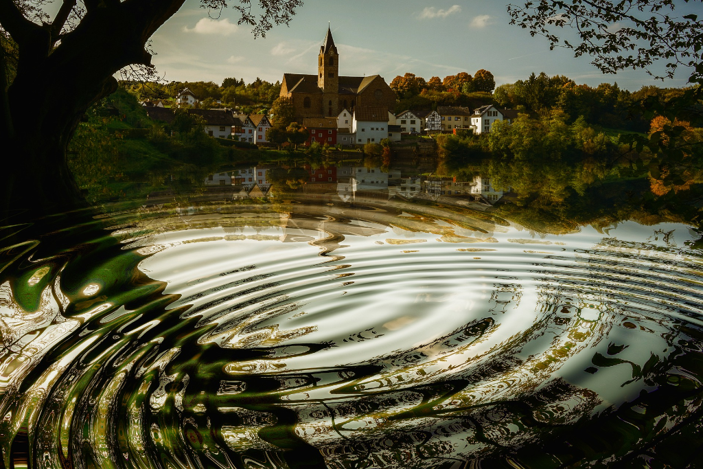 Image of lake with ripples of water stretching out underneath tree limbs and a church visible on the other side of the lake - photo by Gerd Altman on pixabay